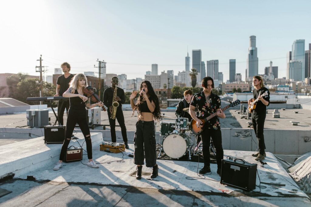 A diverse indie band performs energetically on a rooftop with a stunning Los Angeles skyline backdrop.