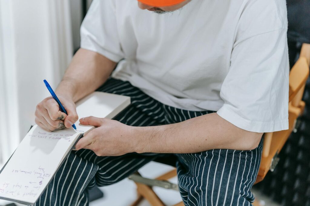 A man sits indoors writing notes with a pen in a notepad. Casual setting.