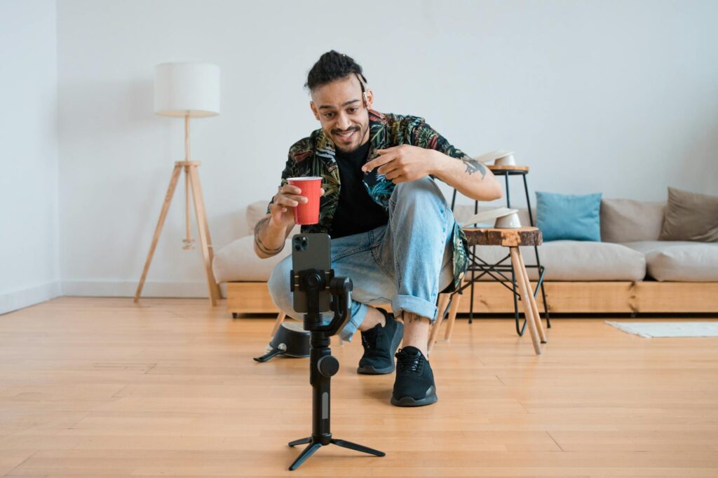 Man filming vlogs in a living room setting, holding a red cup and gesturing at smartphone on tripod.